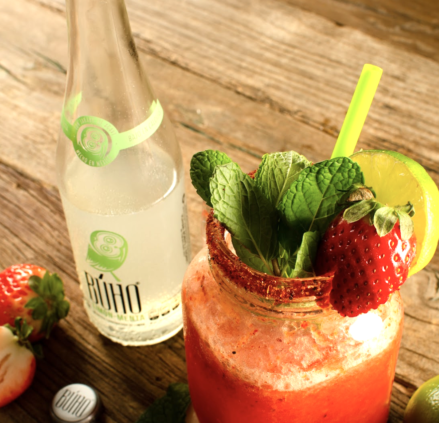 A Bottle of Buho Soda next to a cup with mint, strawberry, and lime
